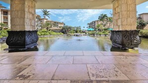 A pool area with two large decorative columns, a clear sky, and surrounding greenery. - H404 - Ocean View Penthouse Suite with AC - Pool, Hot Tub, & Spa - Walk to shops (Kapaa)