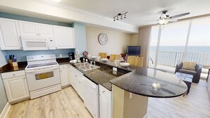 Una cocina moderna con armarios blancos, una estufa y un lavabo. Hay un comedor con una mesa y sillas, y un balcón con vista al océano.