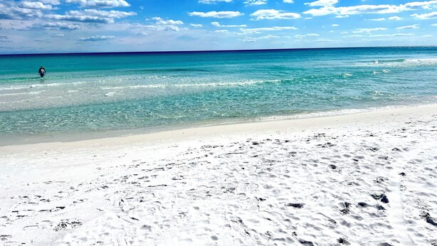 A beach with clear turquoise water, a sandy shore, and a person walking in the distance.
