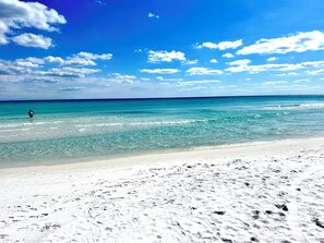 A beach with clear turquoise water, a sandy shore, and a person walking in the distance. - Modern remodeled, spacious 1st floor condo, steps to beaches & restaurants! (Santa Rosa Beach)