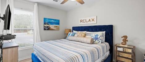 A bedroom with a bed, a ceiling fan, a nightstand, a lamp, a painting, and a sign that says "RELAX".