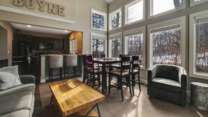 Un vaste espace de vie comprenant une table basse en bois, un canapé gris et une table à manger avec des chaises. Une grande fenêtre offre une vue sur les arbres à l’extérieur.