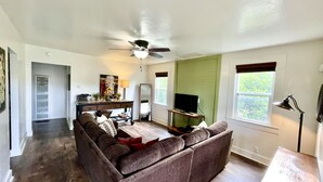 Una sala de estar con un sofá seccional marrón, un ventilador de techo, un televisor sobre un mueble bajo y una ventana con cortinas.