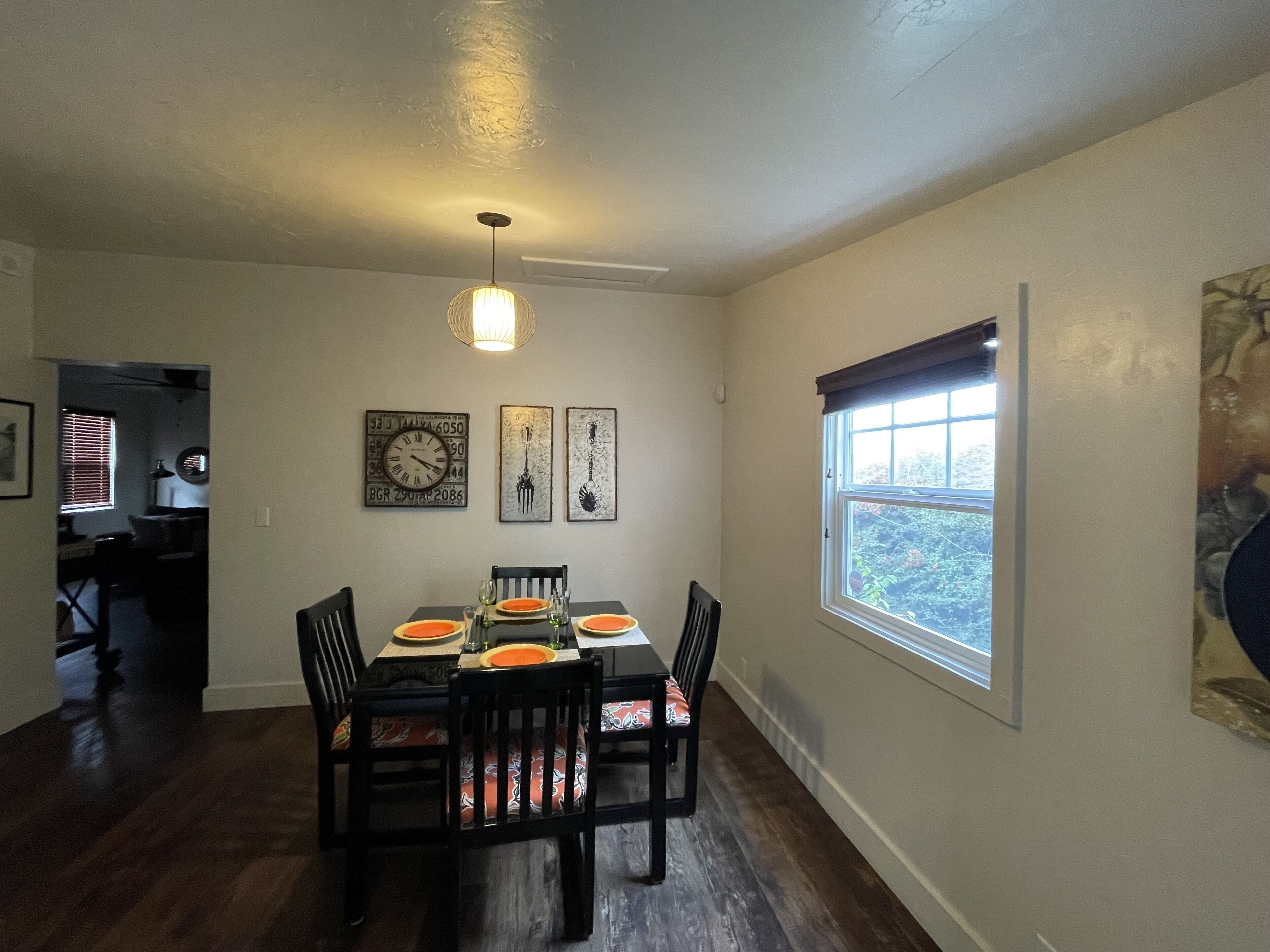 Un coin repas avec une table dressée pour quatre personnes, des chaises avec des coussins orange, une horloge murale et une fenêtre avec des stores.