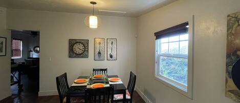 Un coin repas avec une table dressée pour quatre personnes, des chaises avec des coussins orange, une horloge murale et une fenêtre avec des stores.