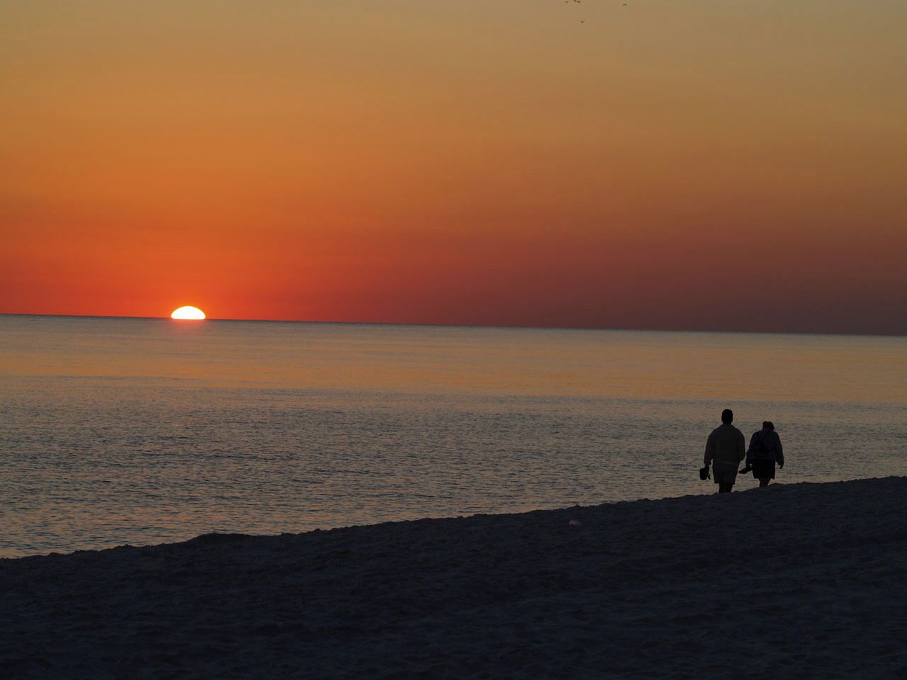 Solnedgang over et roligt hav med to silhuetter, der går langs stranden.