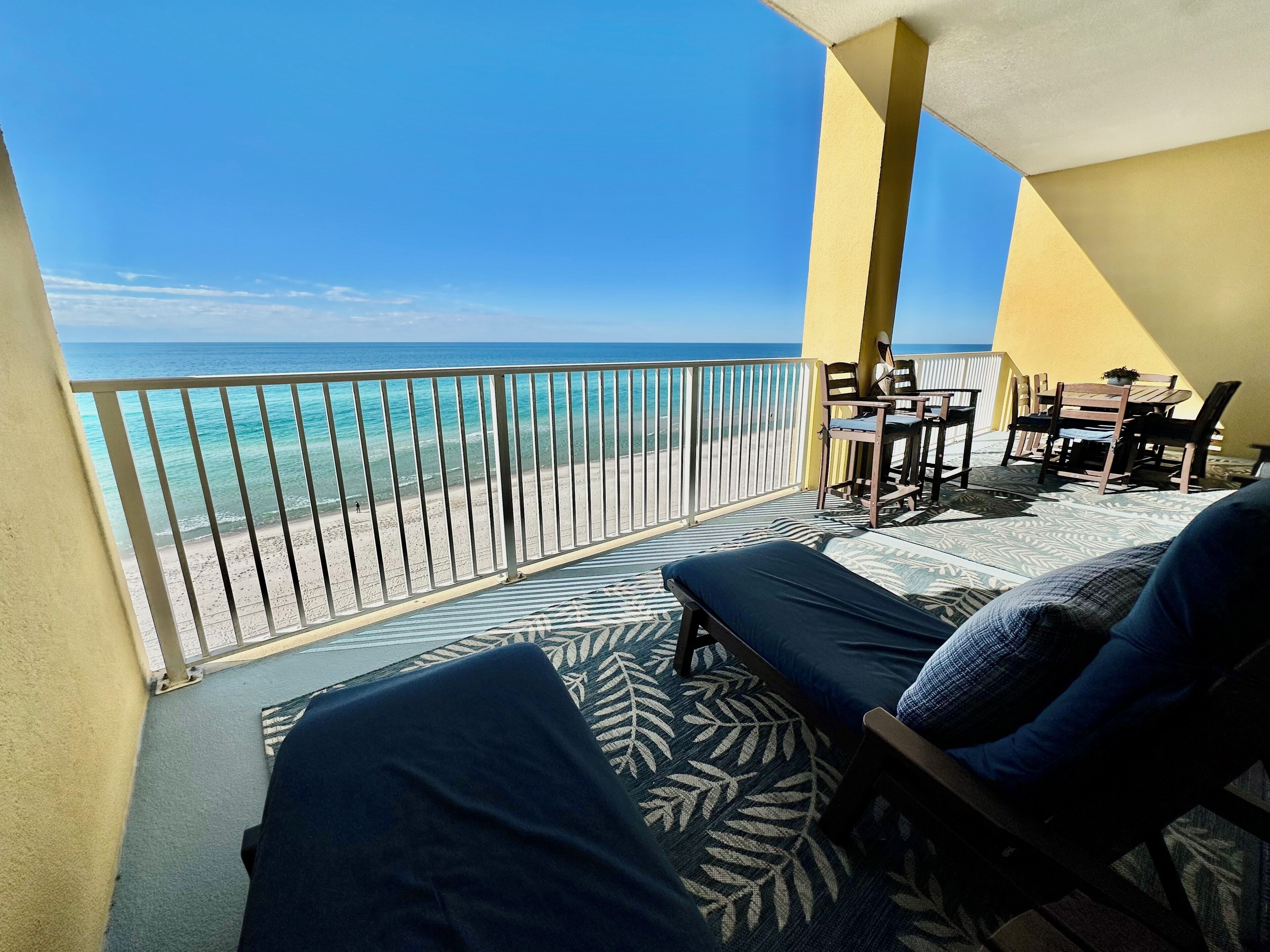 Een balkon met uitzicht op het strand, een tafel met stoelen en een bed met een geruit tapijt.