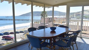 A dining area with a table and chairs, large windows with a view of the beach and pier, and a wooden floor. - Breathtaking Ocean and Beach Views (Avila Beach)
