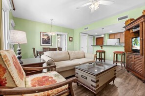 Una sala de estar con sofá, sillón, mesa de centro y televisión. Cuenta con una zona de cocina con armarios y un refrigerador.