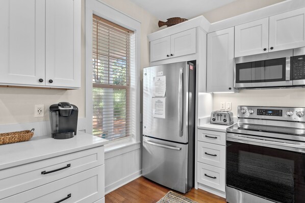 A modern kitchen with white cabinetry, a stainless steel refrigerator, a built-in oven, and a coffee maker. - Charming Beach Cottage w/ 2 GOLF CARTS! Pool, Hot Tub, Beach Tram, Near Wharf (Miramar Beach)