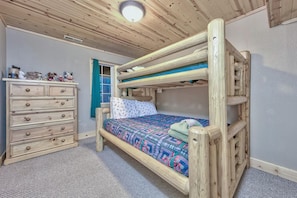 Ein Stockbett mit gemustertem Bettbezug, eine Holzkommode mit Schubladen und ein Fenster mit blauen Vorhängen.