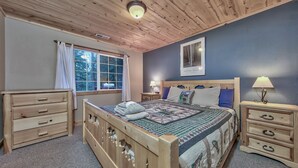 A bedroom with a wooden bed, two bedside tables with lamps, a window with curtains, and a wall-mounted picture.
