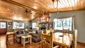 A spacious living area with a wooden ceiling, a fireplace, a dining table with chairs, and a television in the background.