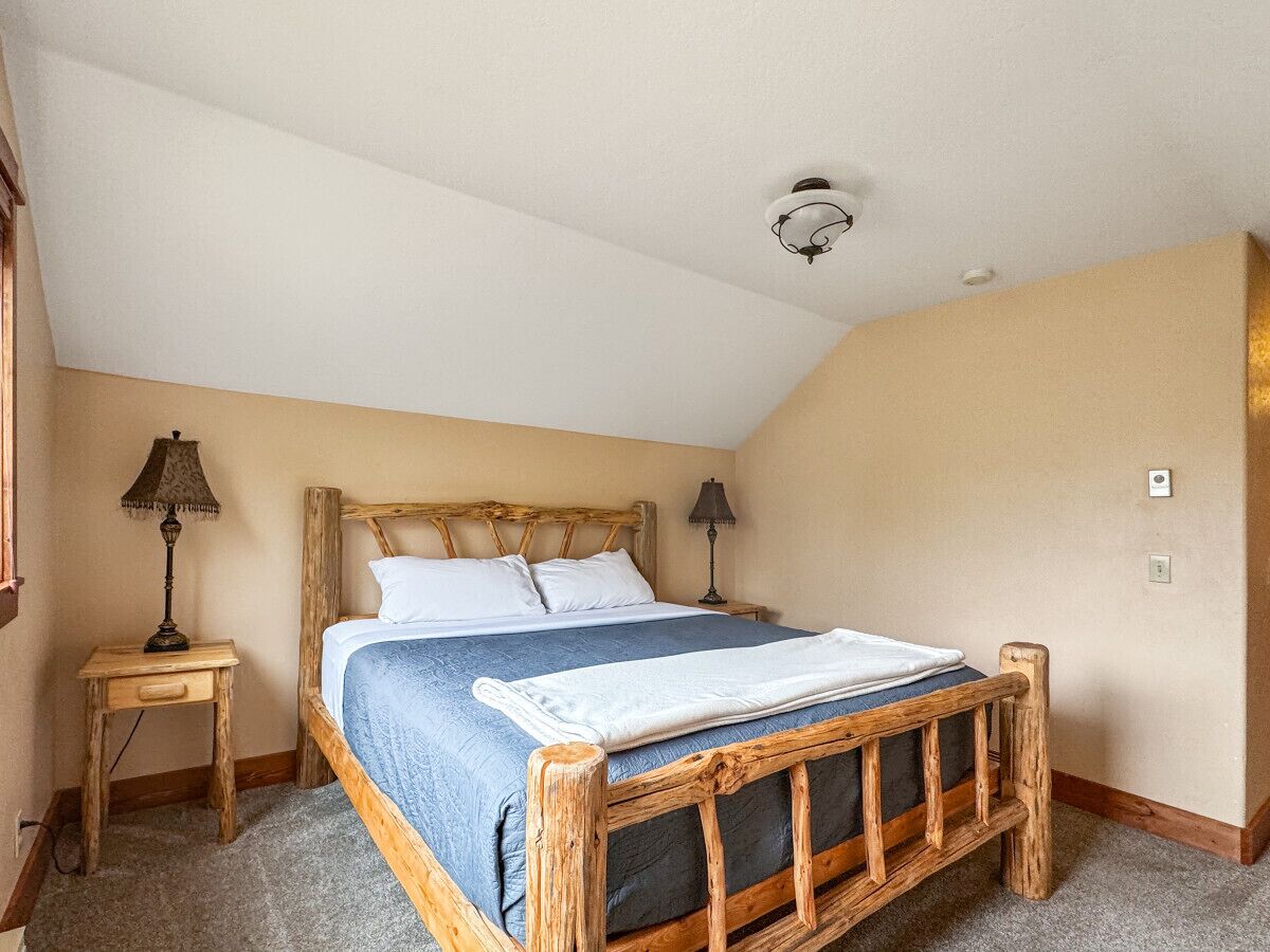 A bedroom with a wooden bed frame, a blue bedspread, two bedside tables with lamps, and a ceiling light fixture.