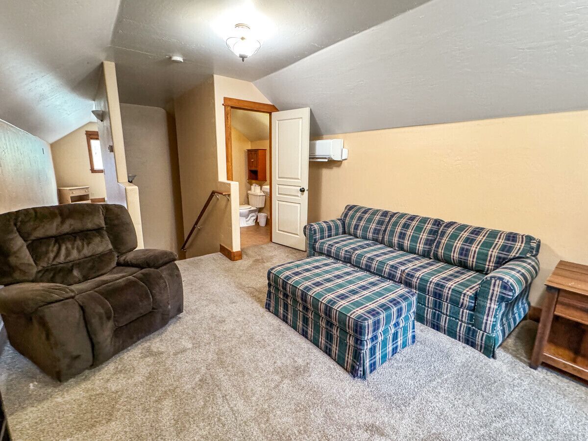 A living room with a brown recliner, a plaid sofa, and matching ottoman. A wooden side table is visible.