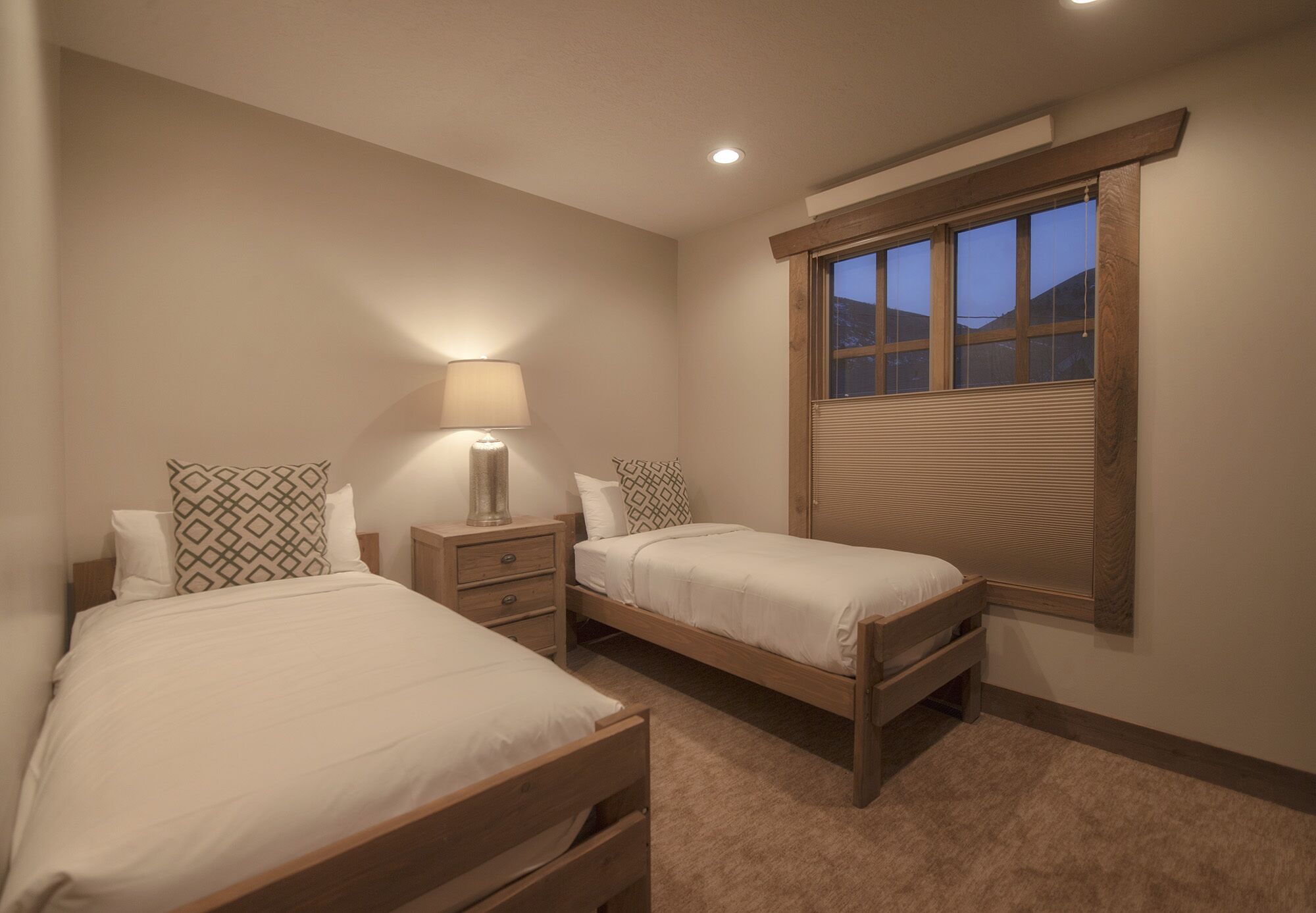 A bedroom with two single beds, a nightstand with a lamp, a window with blinds, and a ceiling light.