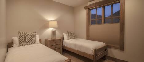 A bedroom with two single beds, a nightstand with a lamp, a window with blinds, and a ceiling light.