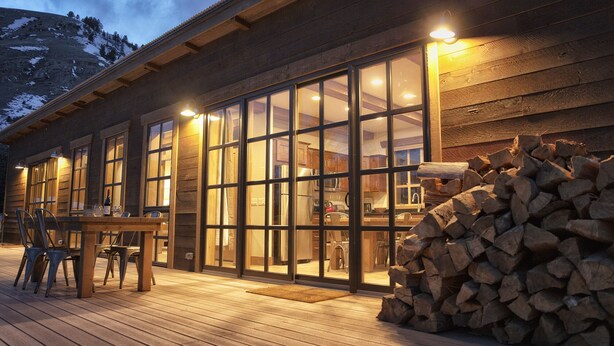 Eine Holzhütte mit großen Glasüren, einer Terrasse mit Stühlen und einem Holzstapel.