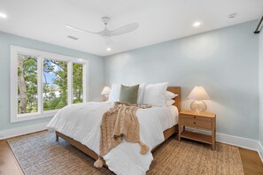 Un dormitorio con cama, mesitas de noche, ventilador de techo y ventana con vista a árboles.