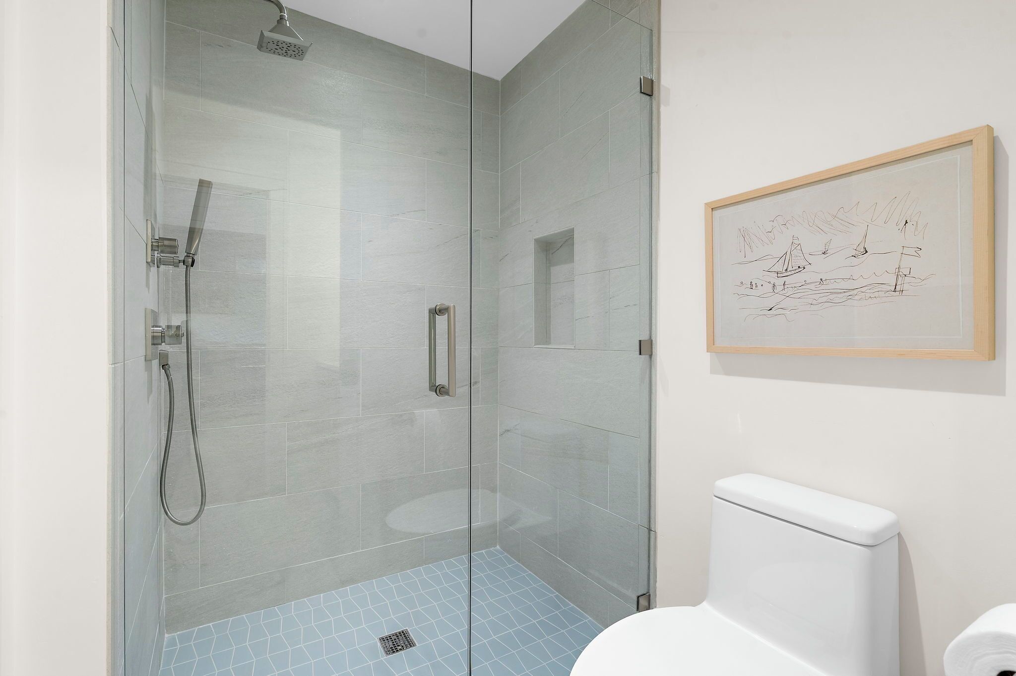A glass-enclosed shower with a handheld showerhead, a toilet, and a framed sketch on the wall.