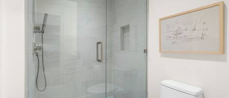 A glass-enclosed shower with a handheld showerhead, a toilet, and a framed sketch on the wall.