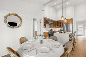 Un coin repas avec une table ronde dressée pour six personnes, entourée de chaises en osier, une cuisine avec des meubles en bois et un grand miroir au mur.
