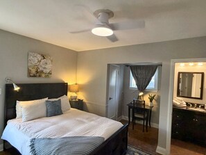 A bedroom with a bed, bedside lamps, a ceiling fan, a desk with a chair, a mirror, and a bathroom with a sink and towel rack.