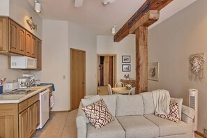 Un espacio habitable compacto con kitchenette, sofá con cojines estampados, armarios de madera y zona de comedor con mesa y sillas.