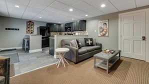 A modern kitchen with dark cabinets, a central island, and a microwave. There is a gray sofa with a cross pillow, a small round table, and a bench with a tray. - 2BR Slopeside Disciples Village 691 - Amazing Ski In/Ski Out on Boyneland! (Boyne Falls)