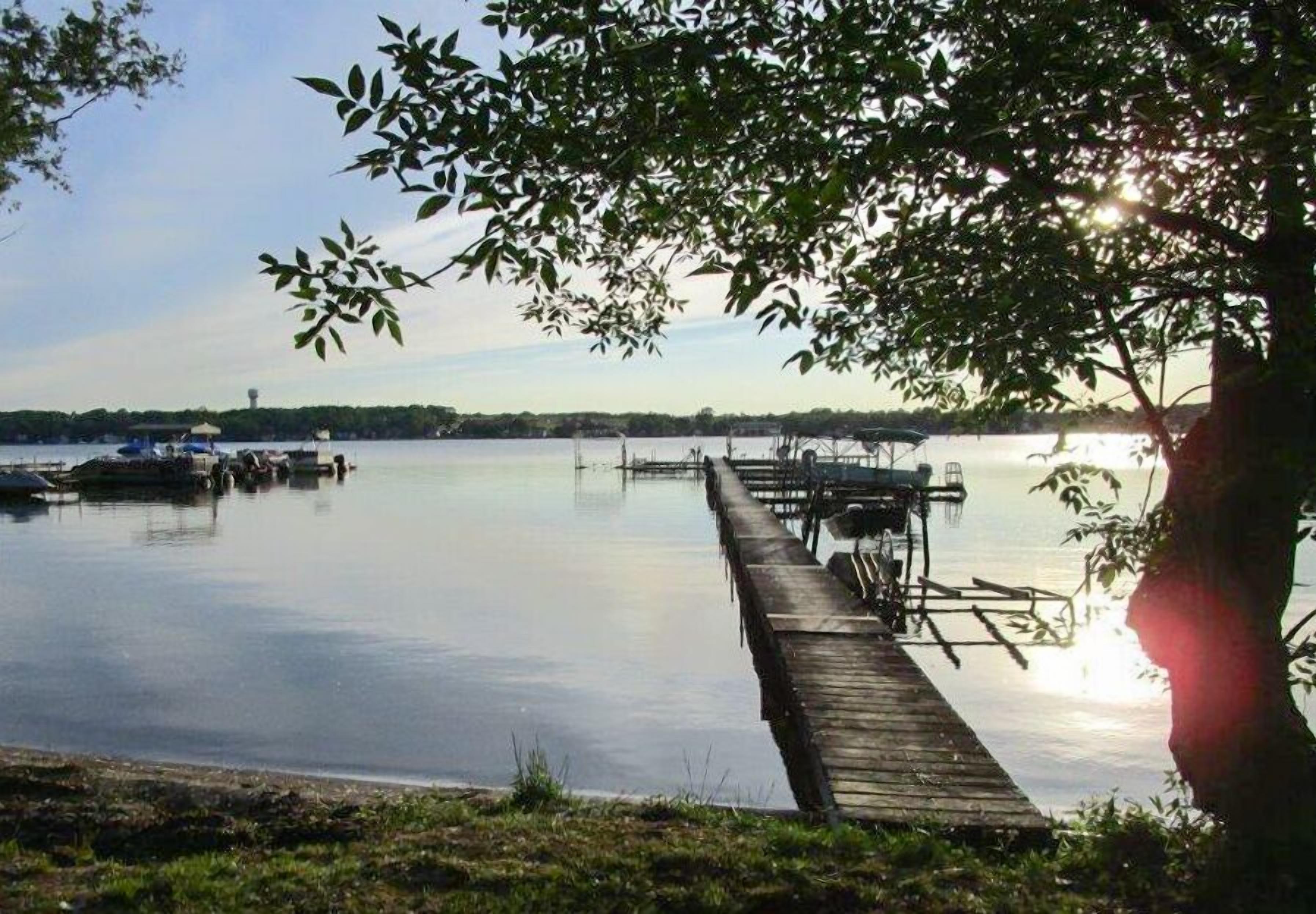 A calm lake with a wooden dock extending into the water, surrounded by trees and boats.