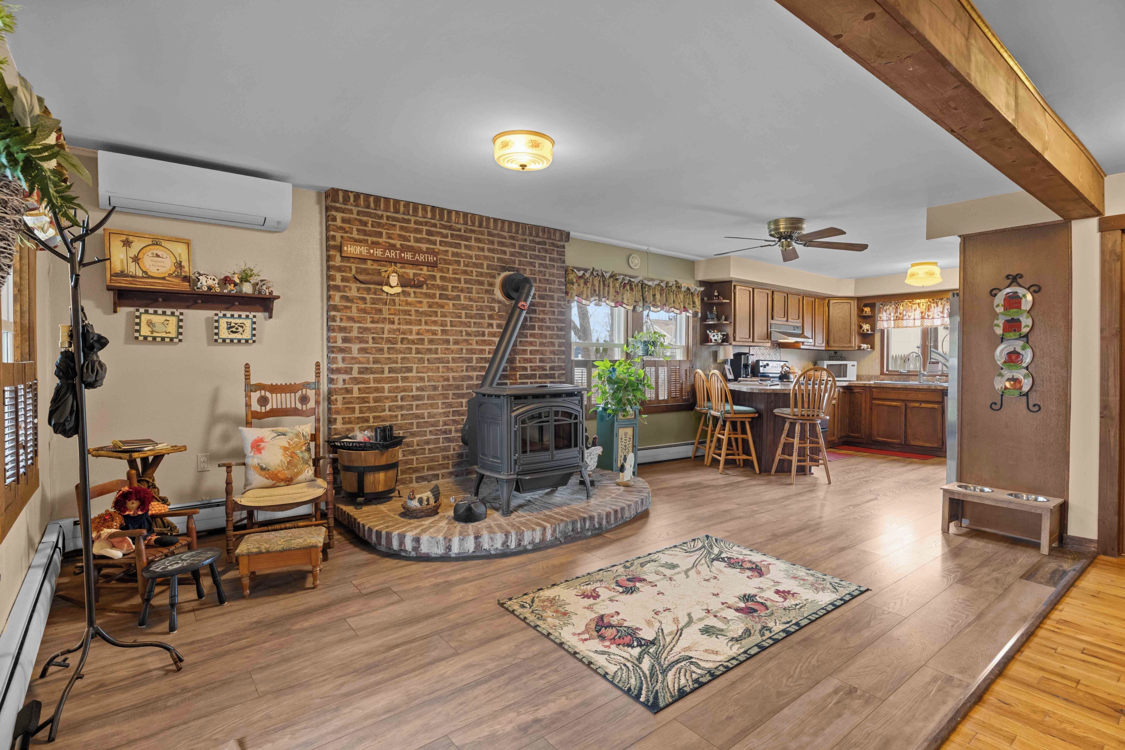 A living room with a fireplace, wooden flooring, a ceiling fan, and a kitchen area with wooden cabinets.