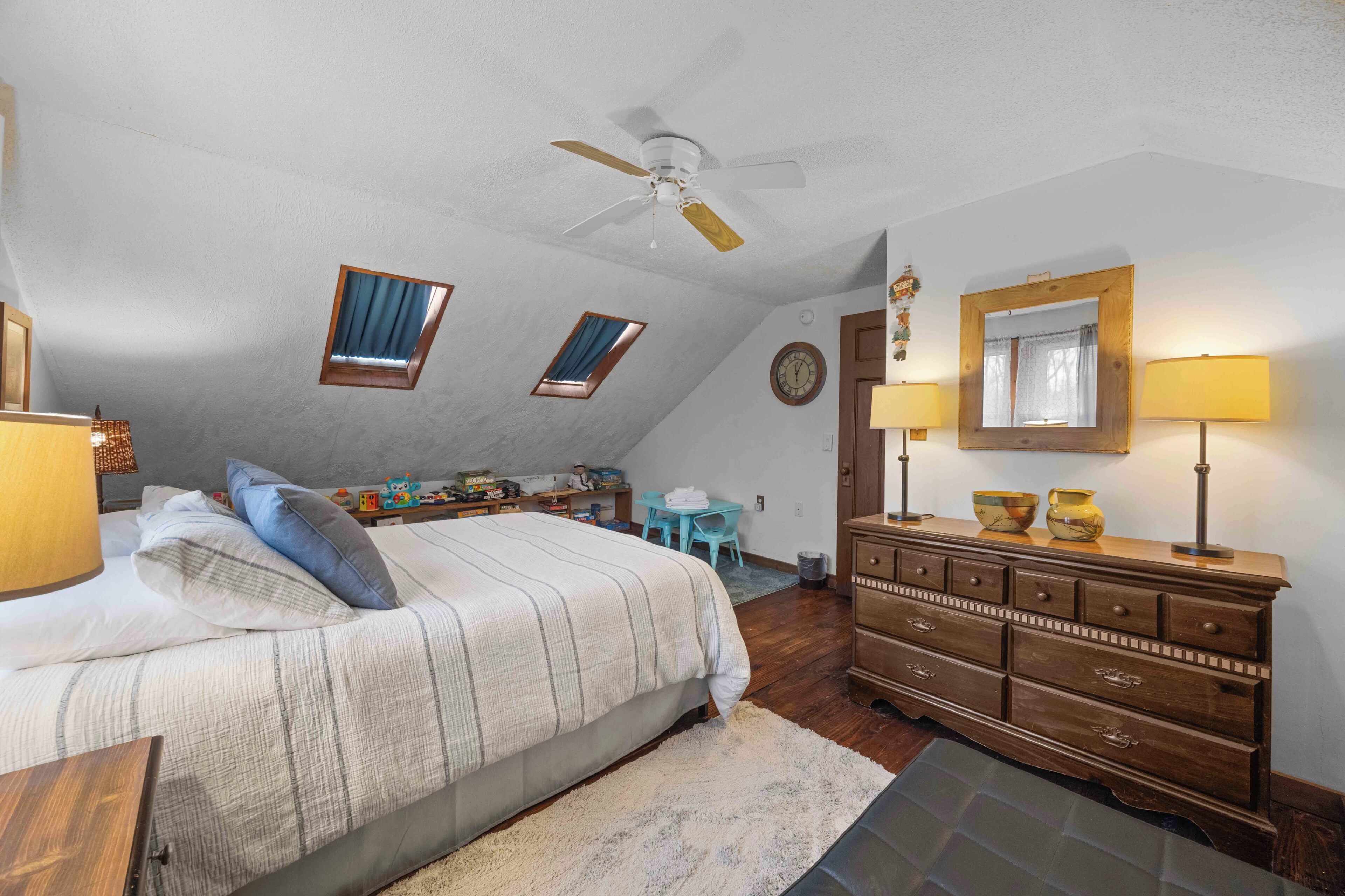 Une chambre à coucher comprenant un lit, une commode, un ventilateur de plafond, deux fenêtres et un bureau avec une chaise.