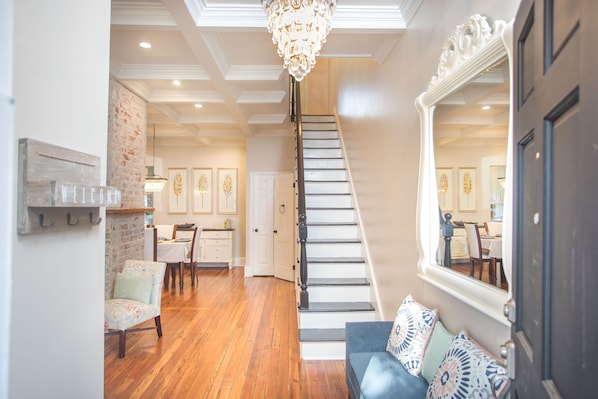 Un escalier menant à une salle à manger agrémentée d’un lustre, d’un canapé avec des coussins à motifs et d’une pancarte « BIENVENUE » accrochée au mur.