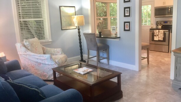 A living room with a blue sofa, a glass coffee table, and a chair. There is a lamp, a picture frame, and a kitchen visible through an open door. - Grace's Getaway: Newly refurbished.Two blocks to the beach! (Jekyll Island)