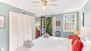 A bedroom with a large bed, white shutters, a ceiling fan, and a view of greenery through a door.