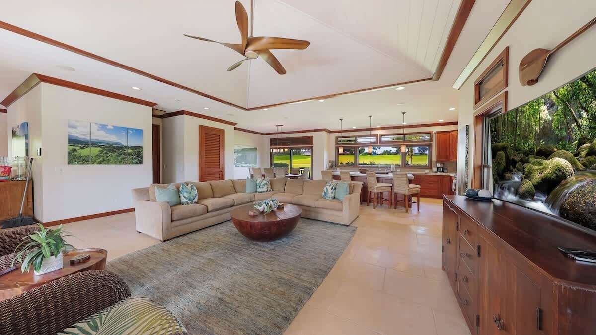 A spacious living room with a large sofa, a coffee table, and a dining area with chairs. There is a large window with a view of a green landscape.
