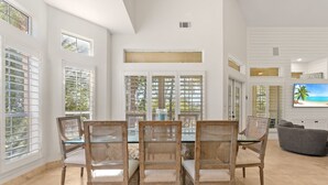 A dining area with a glass-top table surrounded by wicker chairs, large windows with blinds, and a flat-screen TV on the wall. - 4BR Beach Villa with TWO GOLF CARTS!  Near Pool & Beach Tram. Screened Porch. (Miramar Beach)