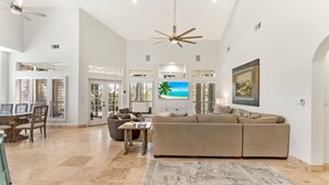 A spacious living area with a large beige sofa, a coffee table, and a TV mounted on the wall. There is a dining area with a table and chairs, and a ceiling fan. - 4BR Beach Villa with TWO GOLF CARTS!  Near Pool & Beach Tram. Screened Porch. (Miramar Beach)