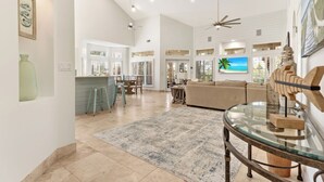 A spacious living area with a high ceiling, a glass coffee table, a beige sofa, and a television mounted on the wall. - 4BR Beach Villa with TWO GOLF CARTS!  Near Pool & Beach Tram. Screened Porch. (Miramar Beach)