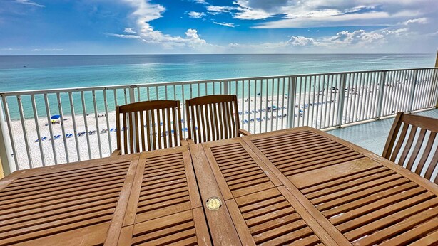 Une table et des chaises en bois sur un balcon donnant sur une plage, avec une eau turquoise et limpide et une rive de sable blanc.