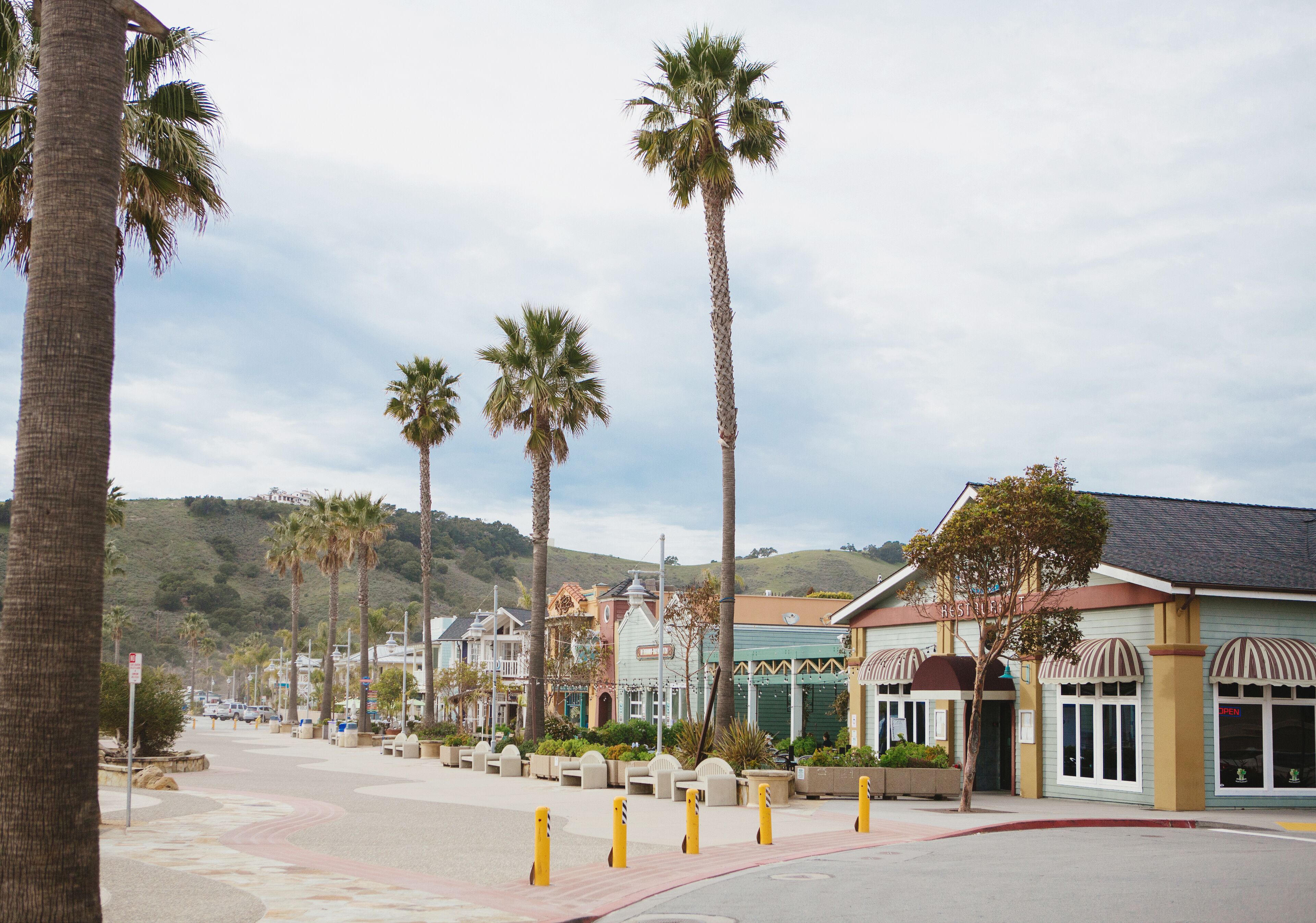 A street lined with palm trees and buildings with a mix of architectural styles, including a building with a sign that reads "OPEN".