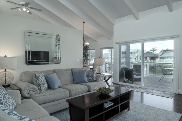 A modern living room with a grey sofa, a coffee table, and a large mirror. There is a balcony with a view of the ocean and buildings. - 4431GB Mandalay Bay Boat Dock & Views Home (Oxnard)