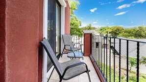 A balcony with two chairs and a table, a red wall, and a view of greenery and a building in the background. - Balcones del Pacifico 21, pool, and Private rooftop access in the heart of town! (Tamarindo)