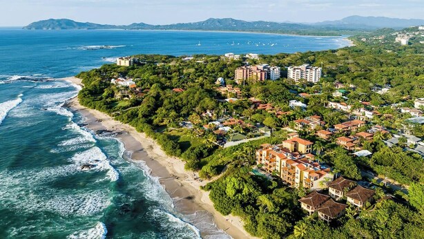 An aerial view of a coastal area with a beach, dense greenery, and residential buildings. - Balcones del Pacifico 21, pool, and Private rooftop access in the heart of town! (Tamarindo)