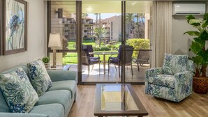 Un salón con sofá, sillón y mesa de centro, una puerta de cristal que da a un balcón con mesa y sillas, y un equipo de aire acondicionado en la pared.