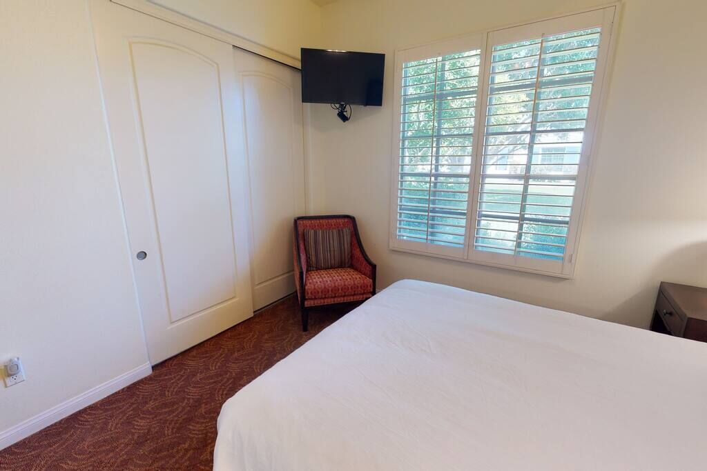 A bedroom with a bed, a chair, a television, and a window with blinds.