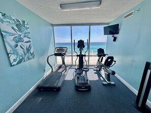 A gym with treadmills and exercise equipment, a large window with a beach view, and a ceiling fan.