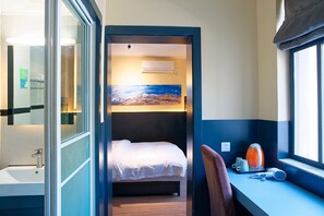 A compact hotel room with a bed, a desk with a chair, a window, and a painting on the wall. - Upland International Youth Hostel (Kunming)