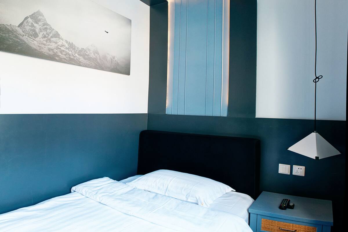 Ein modernes Hotelzimmer mit einem ordentlich bezogenen Bett, einem an der Wand hängenden Bergpanorama und einem schlichten Nachttisch mit Lampe.