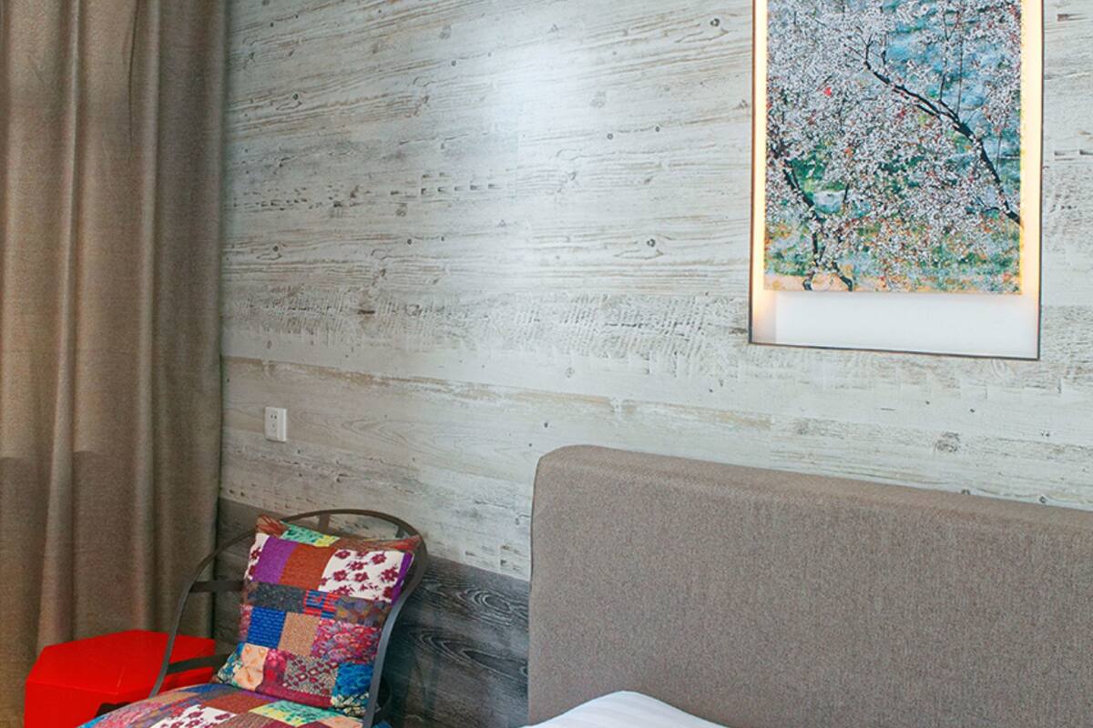 Ein Hotelzimmer mit einem Bett, einem bunten Sessel, einem Kopfkissen und einem gerahmten Kunstwerk an der Wand.
